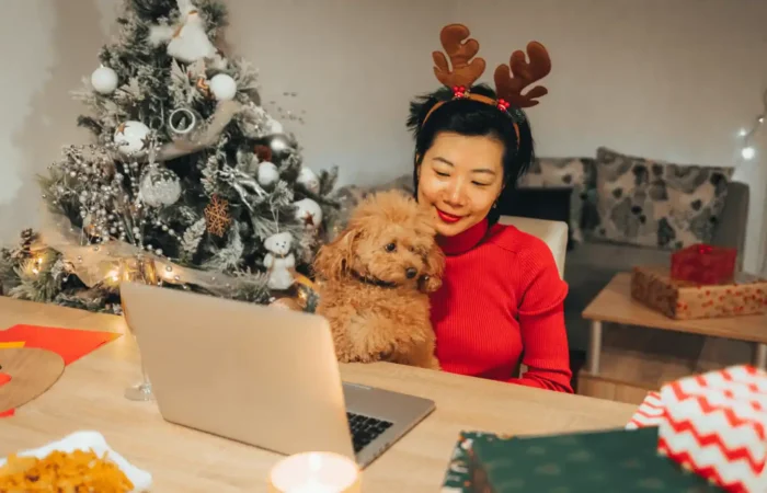 Woman wearing reindeer headband holding her dog while joining a video call beside a decorated Christmas tree