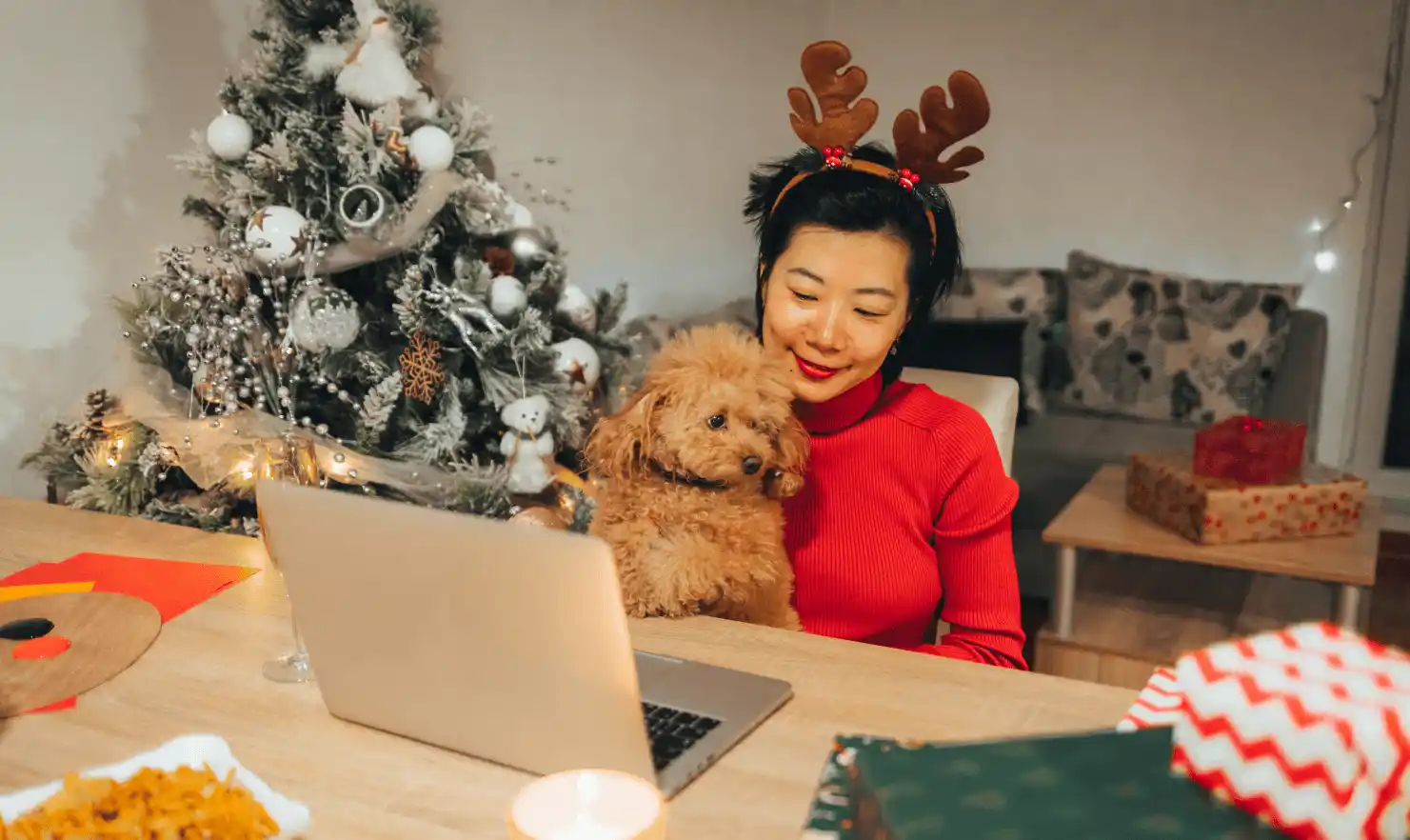 Woman wearing reindeer headband holding her dog while joining a video call beside a decorated Christmas tree