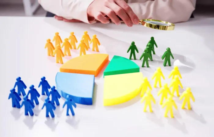 Hand using a magnifying glass to examine colorful figurines grouped around pie chart segments symbolizing market segmentation