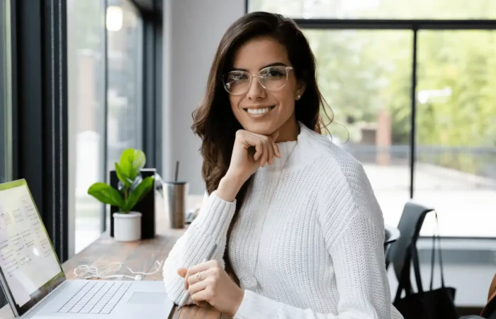Professional woman sitting at a laptop in a bright workspace, smiling confidently while working near large windows