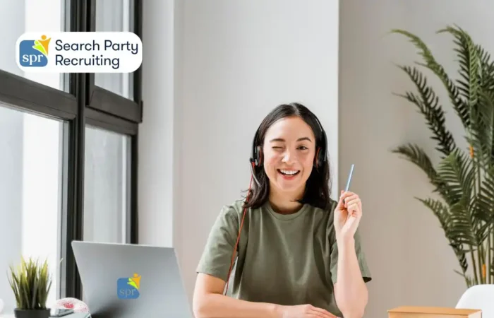 Remote worker smiling and winking while wearing a headset at her desk, shown with Search Party Recruiting branding