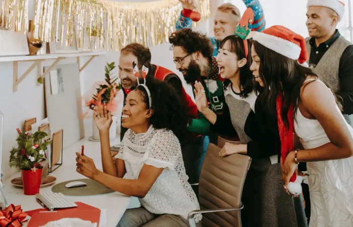 Office team wearing Christmas hats gathered around a computer, celebrating and smiling during a workplace holiday event