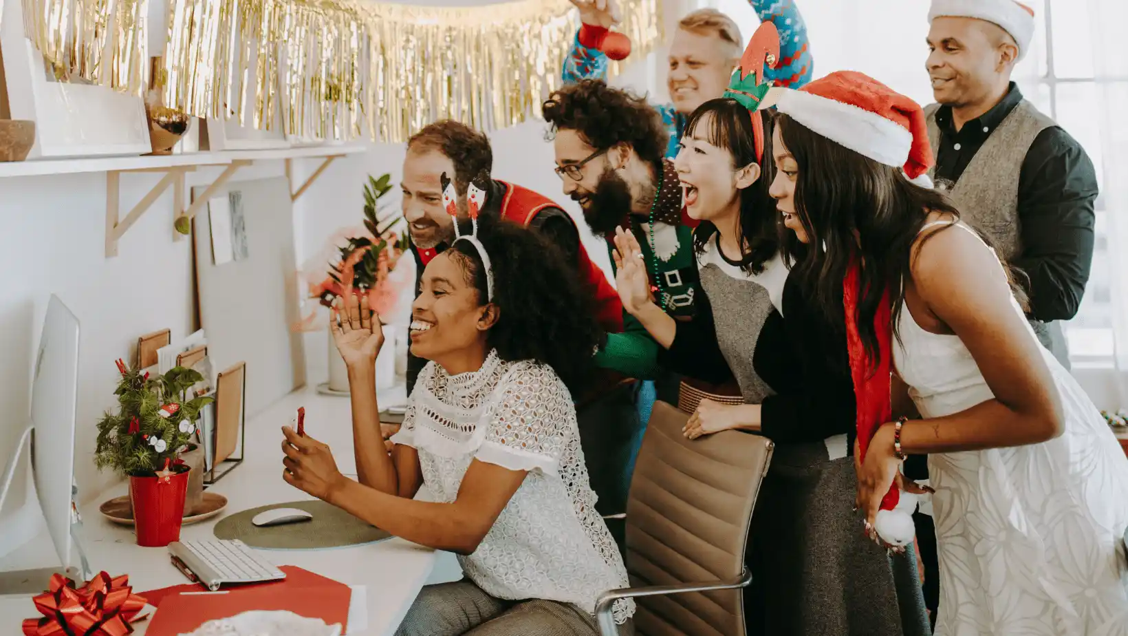 Office team wearing Christmas hats gathered around a computer, celebrating and smiling during a workplace holiday event