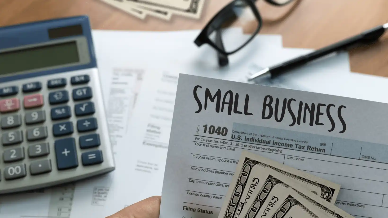 Person holding small business tax form with cash, calculator, and paperwork on a desk